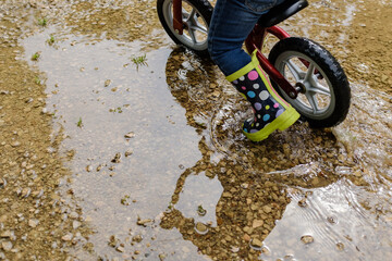 Child playing on water puddle with a toy bicycle and spot rain boots