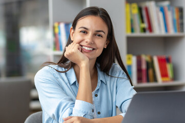 Fototapeta premium Beautiful female student or female employee sitting at table in the university library or office with laptop looking at the camera. Remote work or education concept
