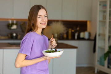 Young and happy caucasian woman eating healthy salad standing in the home kitchen, healthy lifestyle