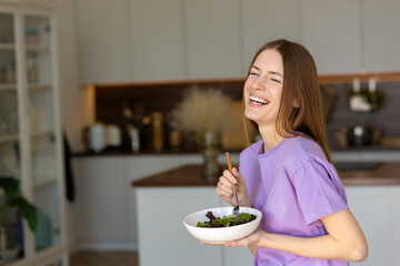 Portrait of young laughing woman with plate of fresh salad standing at home kitchen, happy smiling beautiful girl eating healthy green salad indoors, healthy lifestyle