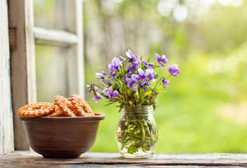 a plate of cookies and a vase of purple flowers stand on the windowsill of an open window against the backdrop of a blurred garden