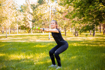 Young attractive woman dressed in sports uniform doing squat plank with dumbbells in hands. Sports activities in the park. Healthy lifestyle concept.
