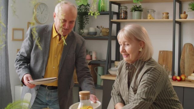 Senior Caucasian woman bringing bottle of homemade lemonade and speaking with husband as he putting plates on dinner table at home