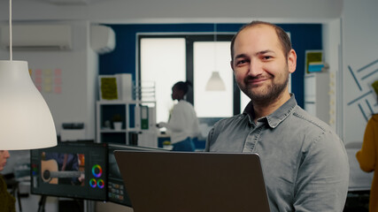 Video editor worker standing in front of camera smiling working in creative agency office holding laptop. Man videographer works in multimedia studio production editing video in modern workplace.