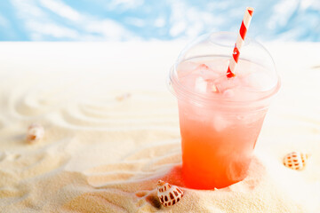 Fresh cold strawberry cocktail in transparent misted plastic glass with straw, ice on sunny beach with ocean and white sand, copy space.