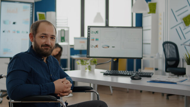 Young disabled paralized handicapped man project manager looking at camera smiling sitting in wheelchair in business office room, working at financial project with team. Businessman with disabilities - Powered by Adobe
