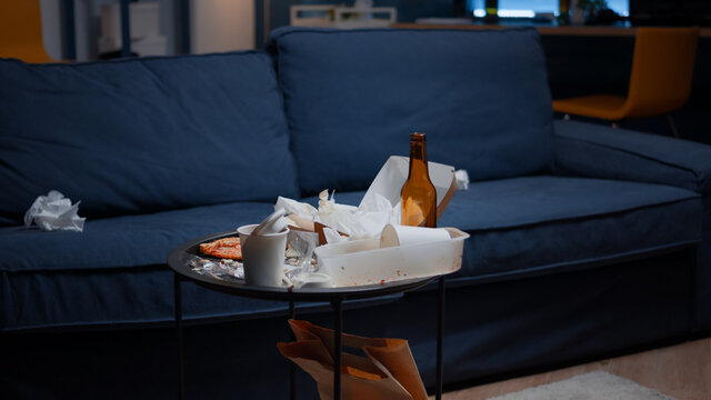 Empty Home Of Depressed Person With Messy Table With Leftover Thrown On Floor In Unorganized Living Room Of Depresive Person. Pizza, Beer Bottles And Napkins In Dirty Apartment