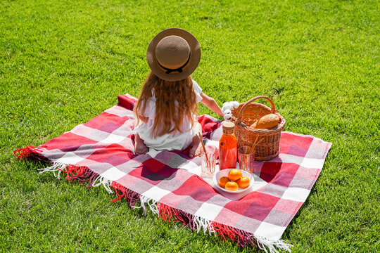 A girl with long hair in a hat sits with her back, a picnic in the park on the green grass at noon, schoolchildren vacation, vacation and travel.