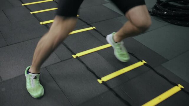 Crop sportsman doing exercises on agility ladder. Unrecognizable active male athlete running on speed ladder and training in gym 