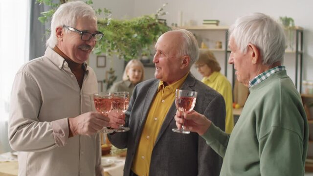Company of three positive senior men clinking glasses together, saying toast and drinking wine at home party with friends