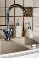 Metallic kitchen faucet, sink from stone and some bottles with soap and cleaner liquids, small...