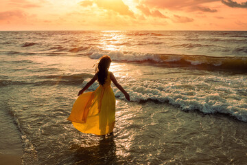 Seascape during golden sunrise with beautiful sky. Woman on the beach. Young happy woman in a yellow fluttering dress walks along the seashore. The girl looks at the magical sunrise. View from above.