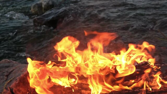 Burning Fire and Embers on Background of River Stones and Stormy River Flows Camping and Recreation by the Water