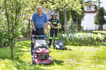 Elderly grandfather teaching her granddaughter how to use mower and cut grass. They work in the rustic garden in th village