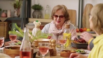 Positive elderly women sitting at dinner table, enjoying meal and speaking with friends at home party