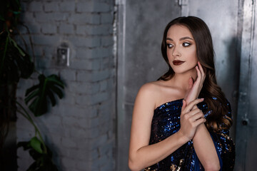Fototapeta premium Close-up portrait of beautiful girl with an evening festive professional make-up in blue, gold dress in sequins on metallic, gray shiny background. Grunge texture of interior. Clean skin, healthy face
