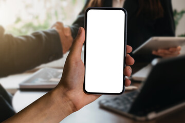 Hand holding white mobile phone with blank white screen in office.