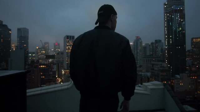 Man standing on the roof of a skyscraper in New York City at night. Back view of man in snapback hat and jacket looking at NYC cityscape from the rooftop