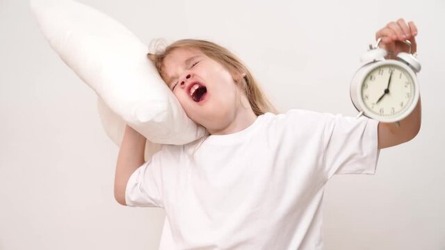A little sleepy girl hugs a pillow and an alarm clock on a white background. early ascents of children to school and kindergarten. comfortable bedding.