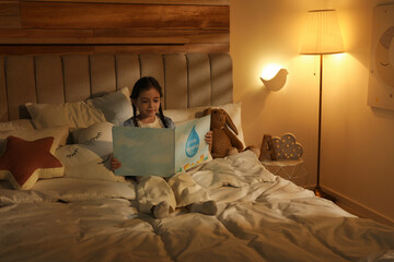 Little girl reading book in bedroom lit by night lamp