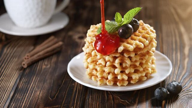 Pouring strawberry syrup on a stack of freshly baked belgian waffles decorated with blueberry and mint on white plate. Delicious homemade pastry for breakfast on wooden table or rustic background
