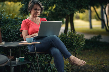 Senior woman sitting in outdoor coffee shop using laptop and drinking coffee.