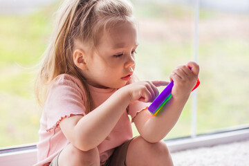 A little blonde girl sits near the window and plays with new trend sensory toy - rainbow pop it. Antistress сolorful toy simple dimple. Squishy soft bubble toys rainbow color