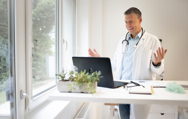 handsome doctor with white coat and blue shirt and stethoscope stands behind high table and works on laptop and has web conference or meeting