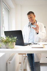 Fototapeta premium handsome doctor with white coat and blue shirt and stethoscope stands behind high table and works on laptop and has web conference or meeting