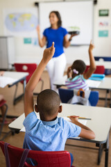 Rear view of african american boy raising his hands while sitting on his desk in the class at school