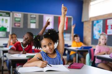 African american girl raising her hands while sitting on her desk in the class at school - Powered by Adobe