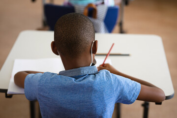 Rear view of african american boy studying while sitting on his desk in the class at school
