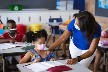 African american female teacher wearing face mask teaching african american girl in class at school