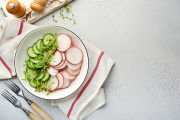 Fresh salad with red radish, cucumber, vegetables, microgreen radishes in white plate on grey stone background. View from above. Concept vegan and healthy eating.