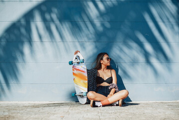 Asian hipster girl in stylish clothing with skateboard sitting near wall