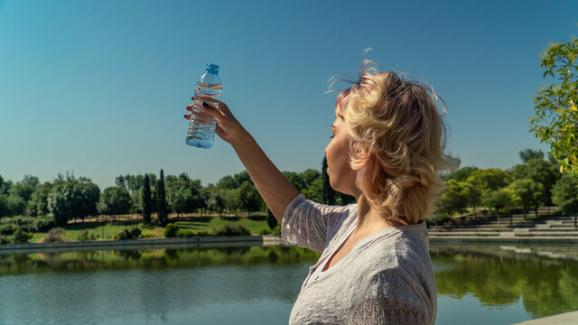 chica joven con los ojos cerrados  levantando una botella de agua al cielo, chica rubia sujetando una botella de plástico al lado del lago