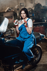 Fototapeta na wymiar Young brunette female mechanic in blue overalls sitting on sportbike in garage or workshop