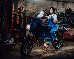 Naklejka na ściany i meble Young brunette female mechanic in blue overalls sitting on sportbike in garage or workshop
