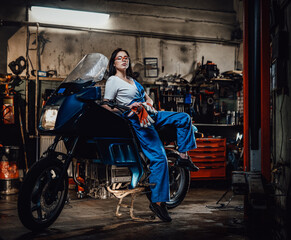 Fototapeta na wymiar Hot brunette girl in blue overalls posing for a camera while leanign on sportbike in garage or workshop