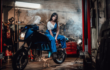 Fototapeta na wymiar Hot brunette girl in blue overalls posing for a camera while leanign on sportbike in garage or workshop