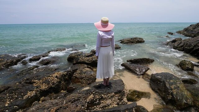 Water slowly move around and splash on stones, woman stand at sea shore and look to small waves. Rough rocks remains at end of sandy beach of Ko Lanta island