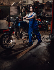Fototapeta na wymiar Beautiful young female mechanic in blue overalls posing with custom bobber in garage or workshop