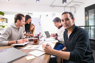 Selective focus creative casual business man confidently looking at camera with background of multiethnic colleagues meeting, brainstorming and discussing ideas with happiness in modern indoor office.