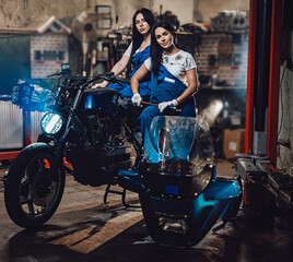Fototapeta na wymiar Two brunette women in blue overalls posing next to a custom bobber in authentic workshop garage