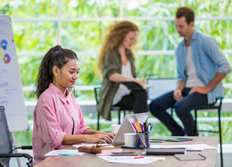 Closeup foreground Asian smart woman typing and using laptop for creative job, smiling with happiness, confidence, comfortably sitting in modern indoor office. Background of multiethnic colleagues.