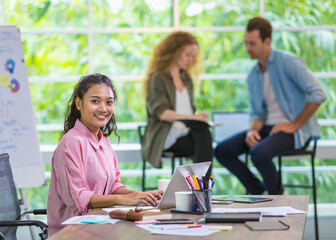 Closeup foreground Asian smart woman typing and using laptop for creative job, smiling with happiness, confidence, comfortably sitting in modern indoor office. Background of multiethnic colleagues.
