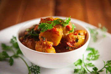 Close-up of Indian vegetarian dish of spicy Potato and Tomato curry garnished with green coriander fresh leaves. Served in a white ceramic bowl over wooden background.