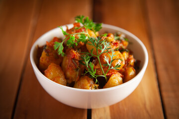 Close-up of Indian vegetarian dish of spicy Potato and Tomato curry garnished with green coriander fresh leaves. Served in a white ceramic bowl over wooden background.