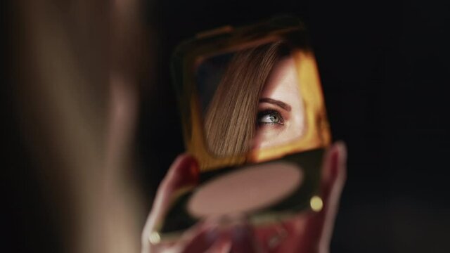Reflection in mirror of womans eyes with shinning make-up. Young lady looks at herself. Makeup at night getting ready before going to party. Slow motion