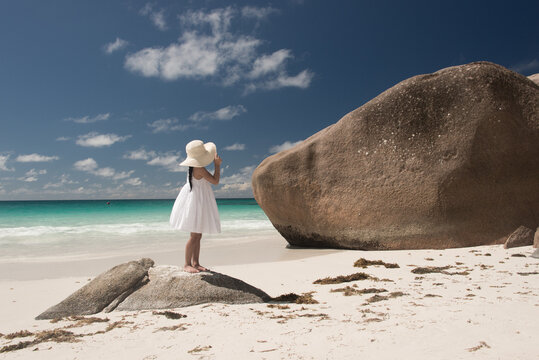 Young girl in white dress and wide hat on tropical beach of Indian Ocean at Seychelles on rock